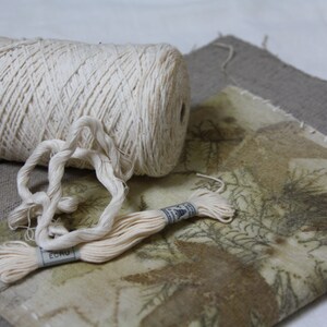 May include: A spool of natural colored cotton thread with a length of thread pulled out and a smaller spool of thread with the word "ECRU" printed on it. The thread is lying on a piece of fabric with a brown and green pattern.