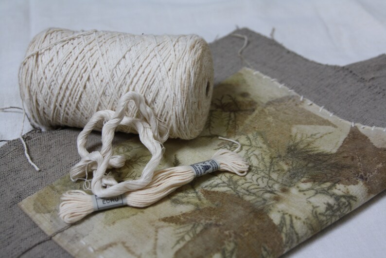 May include: A spool of natural colored cotton thread with a length of thread pulled out and a smaller spool of thread with the word "ECRU" printed on it. The thread is lying on a piece of fabric with a brown and green pattern.