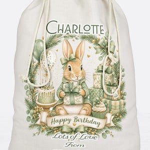 May include: A cream-colored drawstring gift bag featuring a printed illustration of a rabbit holding a gift, surrounded by presents, a cake, and greenery. The text reads Charlotte, Happy Birthday, and Lots of Love From Mum & Dad xxx.