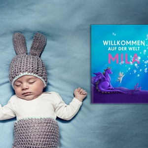 May include: A baby wearing a gray knitted bunny hat sleeps on a blue blanket. A book with the title "Willkommen auf der Welt Mila" is visible in the background.