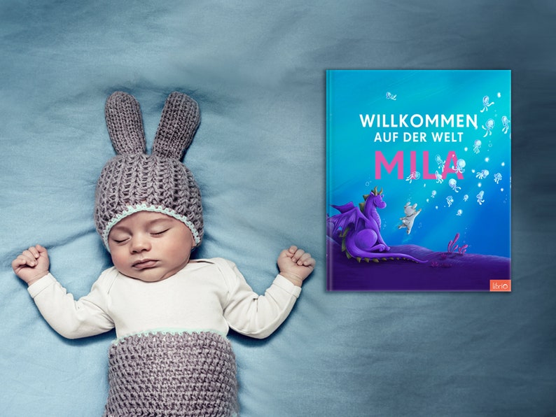 May include: A baby wearing a gray knitted bunny hat sleeps on a blue blanket. A book with the title "Willkommen auf der Welt Mila" is visible in the background.