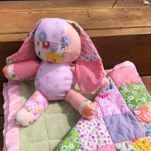 May include: A pink and green patchwork quilt with a pink bunny rabbit toy on top. The quilt is made of different fabrics with floral and butterfly patterns.
