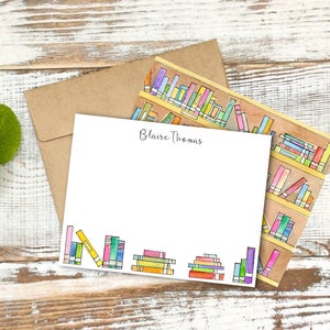 May include: A white card with the name "Blaire Thomas" printed in black cursive script. The card has a border of colorful watercolor illustrations of books on a bookshelf.