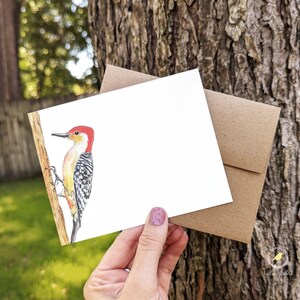 May include: A blank greeting card with a watercolor illustration of a red-headed woodpecker perched on a branch. The card is brown and has a white interior.