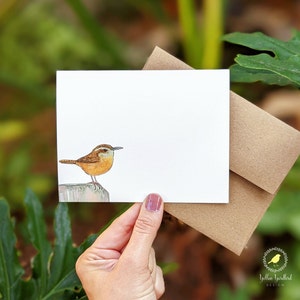 May include: A watercolor illustration of a brown and white bird perched on a wooden post. The bird has a long, thin beak and a small, round head. The background is a white card with a brown envelope.