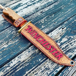 May include: A handcrafted knife with a wooden handle and a leather sheath. The sheath is tan with the word "Winchester" in red lettering. The knife is resting on a weathered, blue wooden surface.