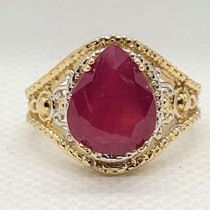 May include: A gold ring with a large, pear-shaped ruby gemstone. The ring has an ornate design with gold and silver accents. The ruby is a deep red color and is the focal point of the ring. The ring is set against a white background.