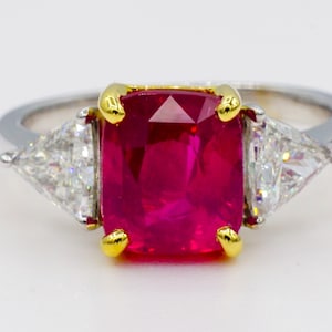 May include: A ring with a large, red, emerald-cut ruby surrounded by two smaller, triangular diamonds.