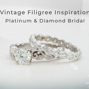 Puede incluir: Dos anillos de novia de platino y diamantes. Uno presenta un gran diamante redondo como piedra central, el otro es una alianza. Los anillos están sobre un fondo blanco. El texto dice: "Vintage Filigree Inspiration Platinum & Diamond Bridal."