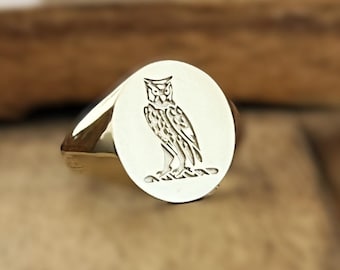 May include: A gold signet ring featuring an oval face with a detailed engraving of an owl perched on a branch. The metal has a polished, reflective finish, and the owl design is centered on the flat surface of the ring, showing clear lines.