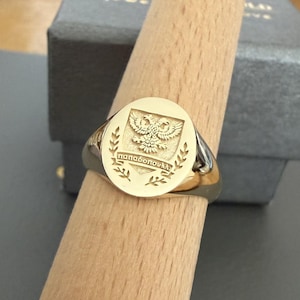 May include: A gold signet ring featuring an engraved double-headed eagle emblem centered on a circular face. Below the eagle, a banner displays the Greek text "παπαδοπουλλ". The ring is displayed on a tapered wooden mandrel for clear viewing.