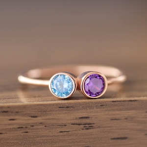 May include: A delicate rose gold ring with two round gemstones. The left stone is a light blue topaz and the right stone is a purple amethyst.