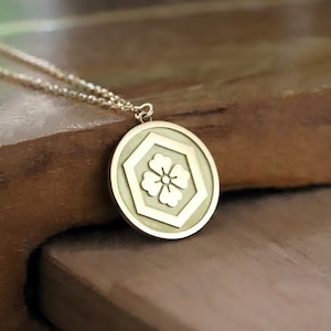 Pode incluir: Um colar de ouro com um pingente redondo que apresenta um desenho de flor branca. O pingente tem formato hexagonal com uma borda elevada.
