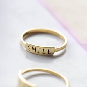 Puede incluir: Un anillo de banda de oro con la palabra "SMILE" grabada en una placa rectangular.