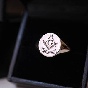 Puede incluir: Un anillo de sello plateado con un emblema mas&oacute;nico, que presenta una br&uacute;jula, una escuadra y la letra "G" dentro de un c&iacute;rculo. El anillo se muestra en una caja negra. El texto "No.9486" est&aacute; grabado en el anillo.