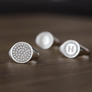 May include: Three silver signet rings with a round face. The ring on the left has a face covered in small, clear stones. The ring in the middle has a face with a single letter "O" engraved in the center. The ring on the right has a face with a single letter "H" engraved in the center.