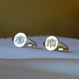 May include: Two gold signet rings with engraved initials. The rings are on a white fabric surface.