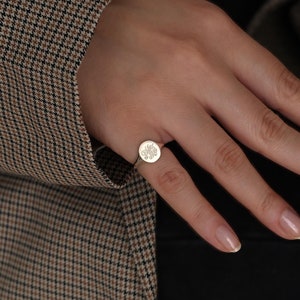 May include: A gold ring with a round, engraved design. The ring is worn on the index finger of a hand.
