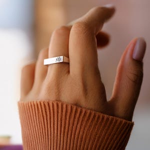 Puede incluir: Un anillo plateado con iniciales grabadas se lleva en un dedo. El anillo es rectangular y tiene un diseño minimalista. La persona lleva un jersey de color óxido.