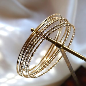 Może przedstawiać: Zestaw pięciu złotych bransoletek, jedna z wzorem skręconej liny, prezentowany na złotej podstawce.