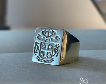May include: A silver signet ring with a square face featuring an engraved coat of arms. The coat of arms includes a crown, a cross, and two lions. The inscription below the coat of arms reads "ANIMO ET FIDE".