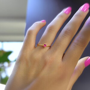 Puede incluir: Un delicado anillo de oro con una pequeña piedra preciosa rosa brillante en forma de corazón. El anillo se muestra en una mano con esmalte de uñas rosa, sobre un fondo borroso. La banda del anillo es fina y elegante.