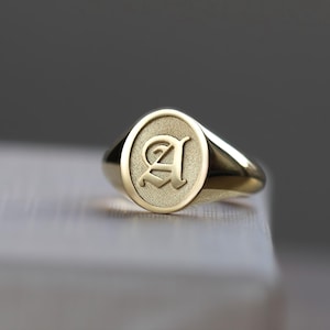 May include: A gold signet ring featuring the letter "A" engraved on its oval face. The ring is made of polished gold and rests on a light surface. The design of the ring is simple and elegant.