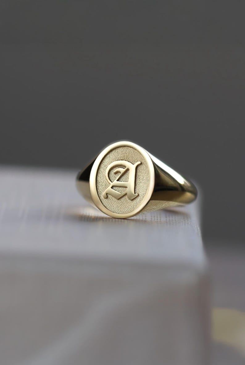 May include: A gold signet ring featuring the letter "A" engraved on its oval face. The ring is made of polished gold and rests on a light surface. The design of the ring is simple and elegant.