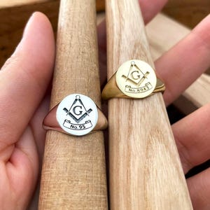 Puede incluir: Dos anillos de sello, uno plateado y otro dorado, cada uno con un s&iacute;mbolo mas&oacute;nico dentro de un dise&ntilde;o circular. El anillo plateado muestra "No. 95", mientras que el dorado muestra "No. 8943". Ambos anillos se exhiben en formas de madera.