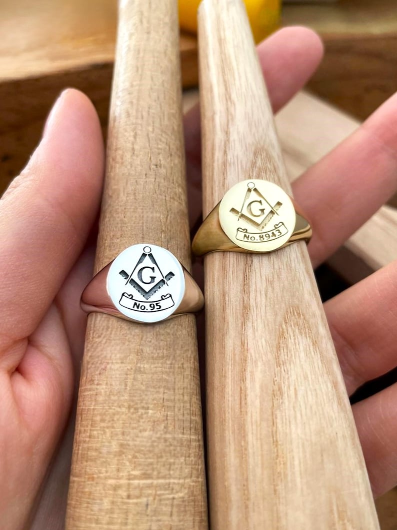 Puede incluir: Dos anillos de sello, uno plateado y otro dorado, cada uno con un s&iacute;mbolo mas&oacute;nico dentro de un dise&ntilde;o circular. El anillo plateado muestra "No. 95", mientras que el dorado muestra "No. 8943". Ambos anillos se exhiben en formas de madera.