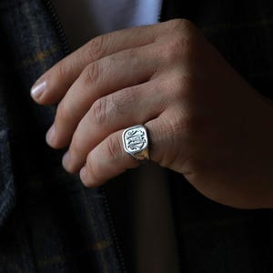 Puede incluir: Un anillo de sello de plata con una cara cuadrada que presenta un diseño grabado. El anillo se lleva en el dedo índice de una mano.