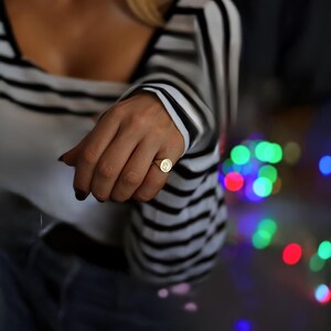 May include: A gold signet ring with a circular face, featuring a stylized letter 'S'. The ring is worn on a finger, set against a backdrop of a black and white striped top and blurred colourful lights.