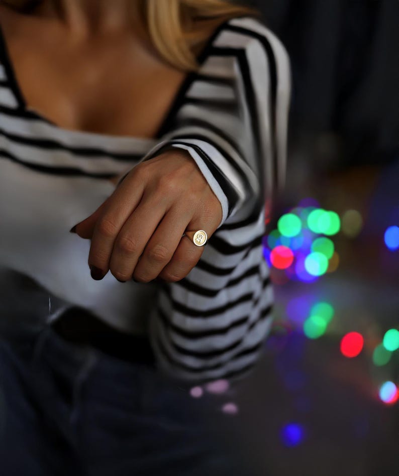 May include: A gold signet ring with a circular face, featuring a stylized letter 'S'. The ring is worn on a finger, set against a backdrop of a black and white striped top and blurred colourful lights.