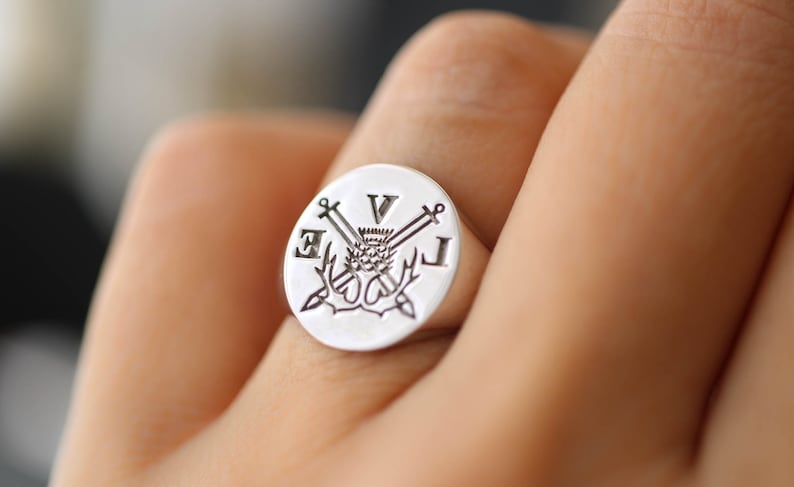 Pu&ograve; includere: Un anello a sigillo in argento con una faccia rotonda che presenta un design inciso di spade incrociate, un cardo e le lettere "V", "E" e "L".