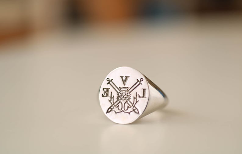 Pu&ograve; includere: Un anello sigillo in argento con una faccia rotonda che presenta un design inciso di un cardo, spade incrociate e le iniziali "E V J".