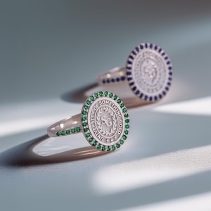 May include: Two silver rings with circular faces. One ring has a green gemstone border and the text "UNIVERSITY OF SOMETHING GOLD". The other ring has a blue gemstone border. Both rings have a detailed emblem in the center.