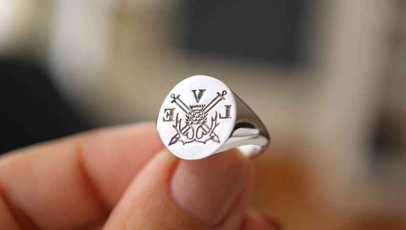 Pu&ograve; includere: Un anello a sigillo in argento con un design inciso che presenta un cardo, spade incrociate e le iniziali "V.J."