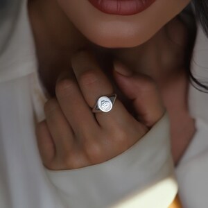 May include: A silver signet ring with a rounded rectangular face. The ring features a raised, stylized initial design. The ring is worn on a finger, with the focus on the ring's design and details.