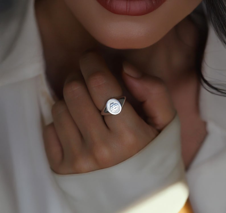 May include: A silver signet ring with a rounded rectangular face. The ring features a raised, stylized initial design. The ring is worn on a finger, with the focus on the ring's design and details.