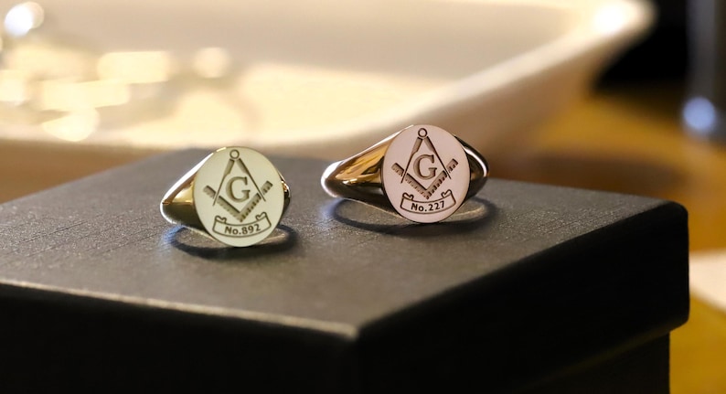 Puede incluir: Dos anillos de sello dorados con un emblema mas&oacute;nico. Cada anillo presenta una cara circular con el s&iacute;mbolo de la escuadra y el comp&aacute;s, la letra "G" y los n&uacute;meros "No. 892" y "No. 227". Los anillos est&aacute;n sobre una superficie negra.