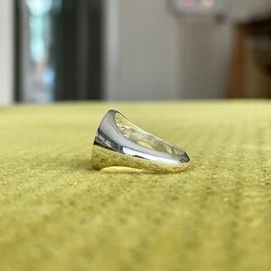 May include: A silver signet ring with a smooth, rounded surface. The ring is designed with a raised edge around the top and a flat band.