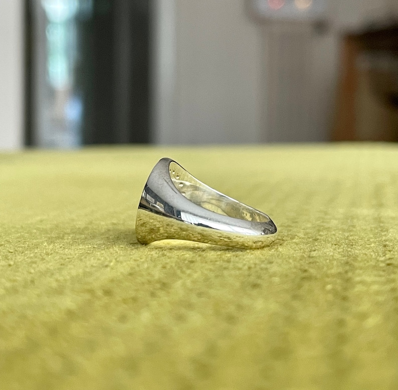 May include: A silver signet ring with a smooth, rounded surface. The ring is designed with a raised edge around the top and a flat band.