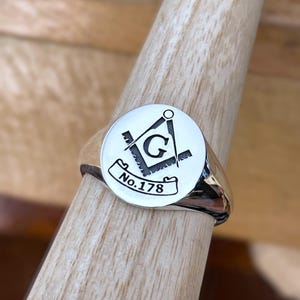 Puede incluir: Un anillo de sello plateado con una cara circular con un s&iacute;mbolo mas&oacute;nico negro y el texto "No.178". El anillo se muestra en una clavija de madera.