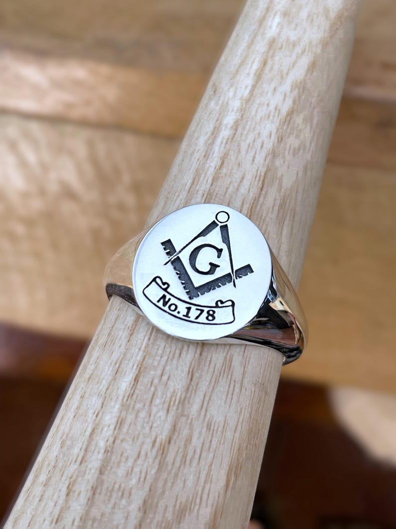 Puede incluir: Un anillo de sello plateado con una cara circular con un s&iacute;mbolo mas&oacute;nico negro y el texto "No.178". El anillo se muestra en una clavija de madera.