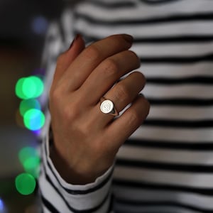 May include: A silver signet ring with a circular face and an engraved design. The ring is worn on a finger, with a striped black and white top in the background. A classic piece of jewellery.