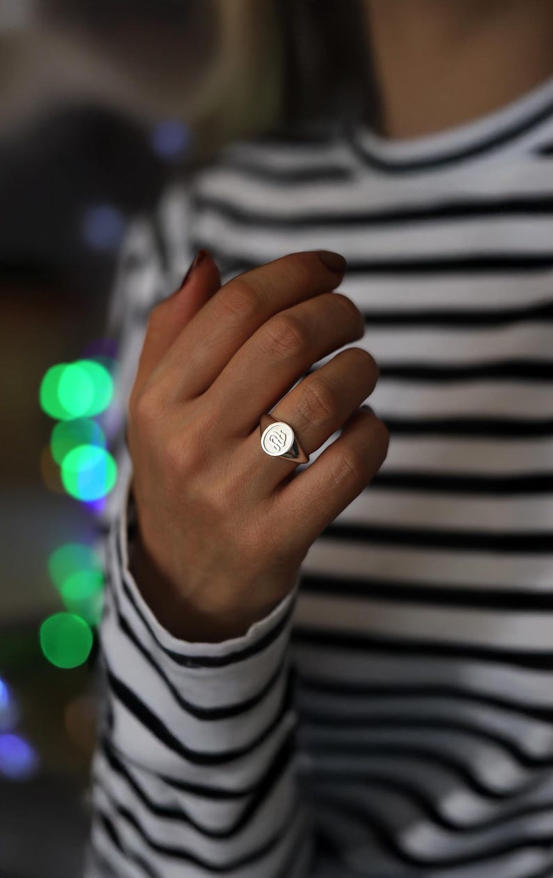 May include: A silver signet ring with a circular face and an engraved design. The ring is worn on a finger, with a striped black and white top in the background. A classic piece of jewellery.