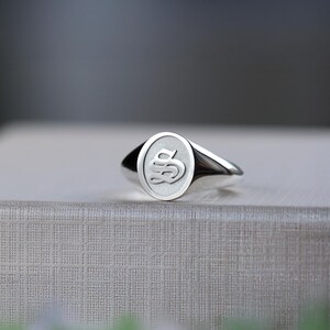 May include: A polished silver signet ring with an oval face. The ring's face displays a raised, stylized letter "S" in a serif font. The ring is highly reflective, with light creating highlights and shadows.