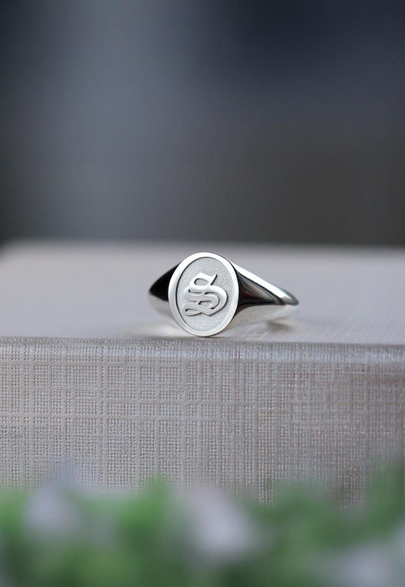May include: A polished silver signet ring with an oval face. The ring's face displays a raised, stylized letter "S" in a serif font. The ring is highly reflective, with light creating highlights and shadows.