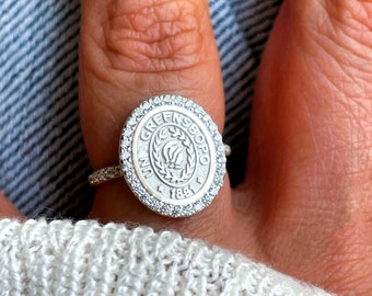 Puede incluir: Un anillo de plata con un diseño circular que presenta las palabras "University of Greensboro" y el año "1891". El anillo está adornado con pequeñas piedras transparentes alrededor del borde.