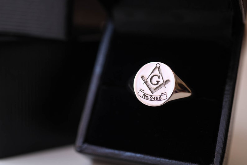 Puede incluir: Un anillo de sello plateado con un emblema mas&oacute;nico, que presenta una br&uacute;jula, una escuadra y la letra "G" dentro de un c&iacute;rculo. El anillo se muestra en una caja negra. El texto "No.9486" est&aacute; grabado en el anillo.